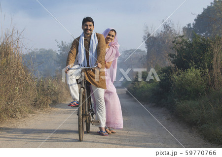 Happy rural couple in traditional dress riding on bicycle on a narrow pathway in a village. 59770766