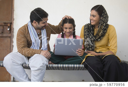Happy village girl sitting with her parents on a cot operating a laptop computer. 59770956