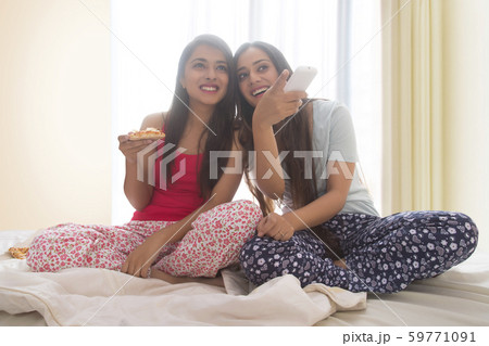 Two smiling young girls sitting on bed at home eating pizza and watching television 59771091