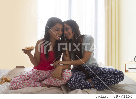 Two young girls sitting on bed at home eating pizza and laughing holding each other Two young girls sitting on bed at home eating pizza and laughing holding each other 59771150