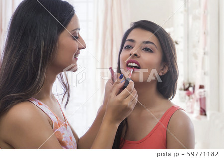 Close up of a smiling teenage girl applying lip paint to her friend at home 59771182