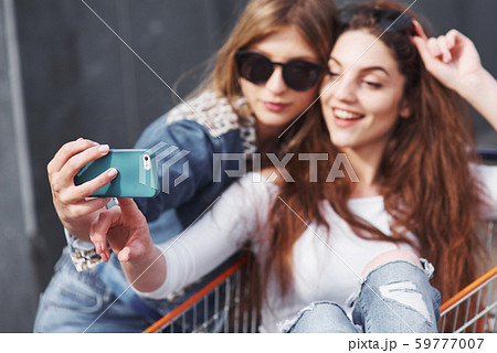 Two young happy women in sunglasses having fun shopping trolley race outdoors. Two young happy women in sunglasses having fun shopping trolley race outdoors. 59777007