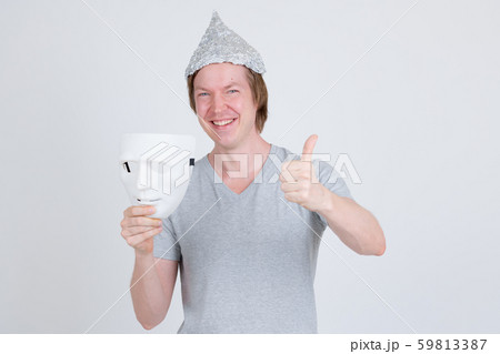 Happy young man with tinfoil hat holding white mask and giving thumbs up Happy young man with tinfoil hat holding white mask and giving thumbs up 59813387