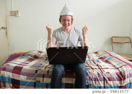Happy young man with tin foil hat using laptop and getting good news in the bedroom 59813577