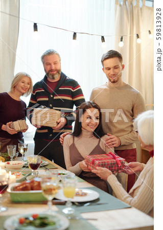 Young affectionate couple giving Christmas present in giftbox to granny by table 59813928