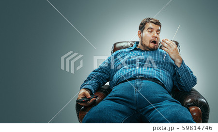 Fat man sitting in a brown armchair, emotional watching TV 59814746