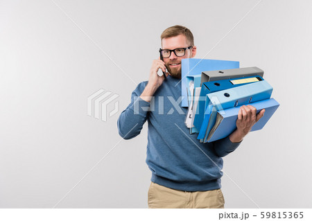 Busy young bearded manager with stack of folders holding smartphone by ear Busy young bearded manager with stack of folders holding smartphone by ear 59815365