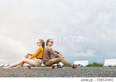 An attractive young couple in sunglasses sit back to back on their longboard in a suburban parking 59852093