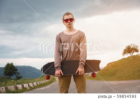 Stylish young man standing along a winding mountain road with a skate or longboard on his shoulder 59854593