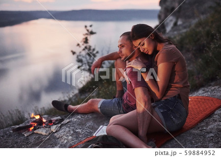 Hugging couple with backpack sitting near the fire on top of mountain enjoying view coast a river or 59884952