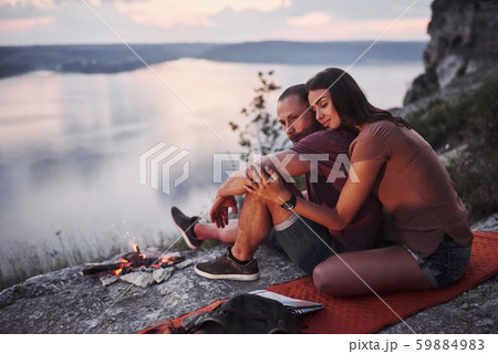 Hugging couple with backpack sitting near the fire on top of mountain enjoying view coast a river or 59884983