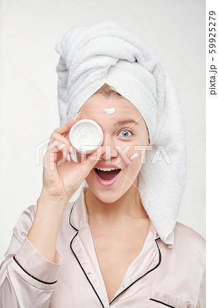 Pretty young ecstatic female with towel on head holding jar of facial cream 59925279