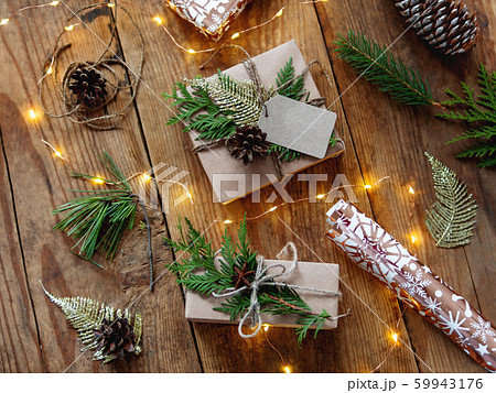 Christmas presents wrapped in craft paper with natural fir tree and thuja branches as decoration. Wooden table with light bulbs and hand made New Year gifts. Christmas presents wrapped in craft paper with natural fir tree and thuja branches as decoration. Wooden table with light bulbs and hand made New Year gifts. 59943176