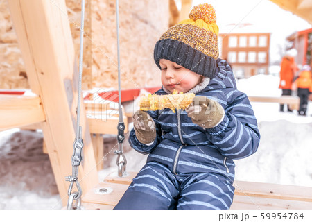 Little boy 3-5 years old, sits on a wooden swing in the winter on the street, eats boiled corn, in a warm hat and blue overalls, background of snowdrifts of snow. Snack for lunch outdoors. 59954784