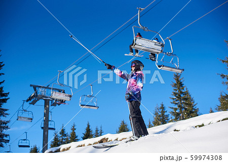 Happy woman skier standing near ski lift on snow-covered mountain slope. Sunny day during winter vacation. General view. Happy woman skier standing near ski lift on snow-covered mountain slope. Sunny day during winter vacation. General view. 59974308