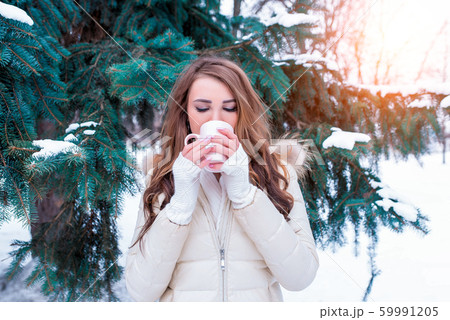 Girl in winter drinks a hot drink from a mug, weekend at resort, background green trees snowdrifts. In a warm jacket, cuffs and a scarf. Heated coffee tea in cold weather. 59991205