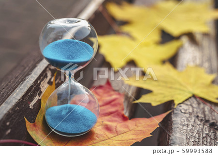 In autumn park on a bench with leaves is hourglass with blu sand. Autumn composition. Transience of time concept. Horizontal image 59993588