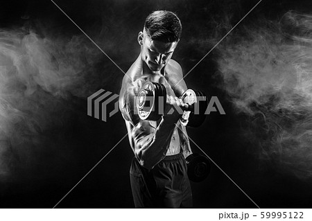 Handsome bodybuilder doing exercise with dumbbell. Studio shot. Black and white photo. Smoke 59995122