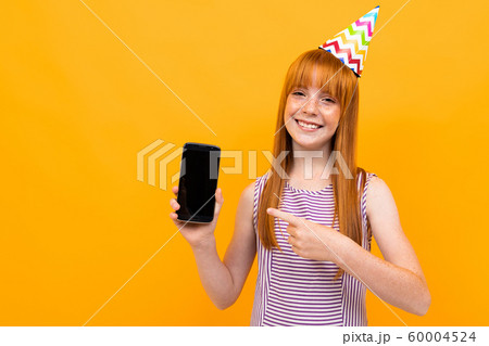 joyful european red-haired girl shows the phone screen over yellow background 60004524
