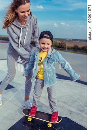 Young mother with little boy son, happy family learns to ride skateboard, summer outdoors, autumn day. Emotions of delight, joy of happiness and fun. Caring support assistance in training, parenting. 60004671