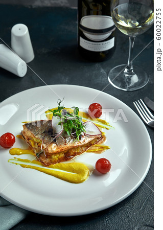 White steak of grilled fish on pillow of vegetables and yellow sauce, with tomatoes and fresh herbs. Fish on white plate with white wine on dark beautiful background. Close-up. Space 60022755