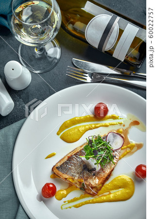 White steak of grilled fish on pillow of vegetables and yellow sauce, with tomatoes and fresh herbs. Fish on white plate with white wine on dark beautiful background. Close-up. Space White steak of grilled fish on pillow of vegetables and yellow sauce, with tomatoes and fresh herbs. Fish on white plate with white wine on dark beautiful background. Close-up. Space 60022757