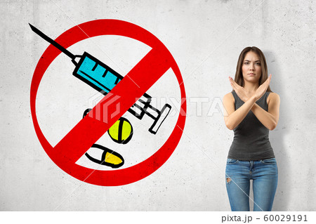 Serious young girl with hands crossed, standing against wall with picture of syringe and some meds enclosed in red prohibition sign. 60029191