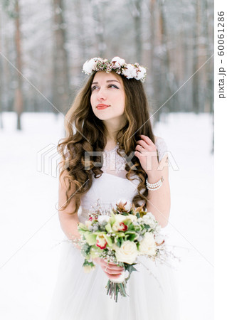 Winter wedding portrait. Portrait of beautiful bride with floral and cotton wreath on the head, holding bouquet, wearing a white dress against the background of the winter forest in snowy weather Winter wedding portrait. Portrait of beautiful bride with floral and cotton wreath on the head, holding bouquet, wearing a white dress against the background of the winter forest in snowy weather 60061286