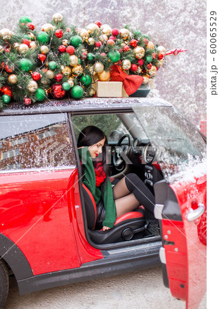 Pretty young girl is sitting in red car with decorated xmas tree on the roof. 60065529
