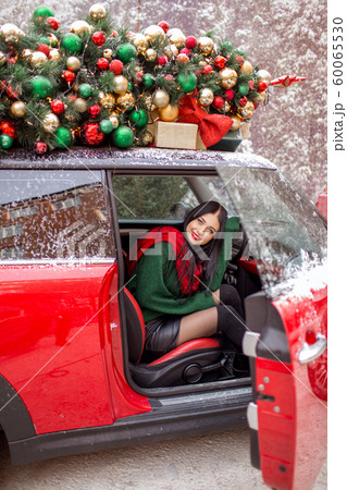 Pretty young girl is sitting in red car with decorated xmas tree on the roof. 60065530