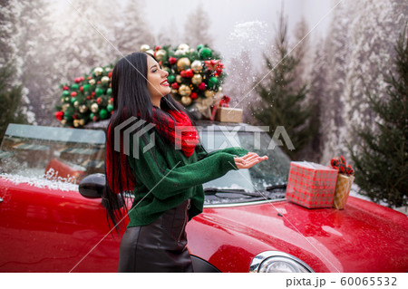 Pretty young girl is playing with snow near red car with decorated xmas tree. 60065532