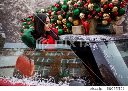 Pretty young girl is standing near red car with decorated xmas tree on the roof, holiday concept. 60065591