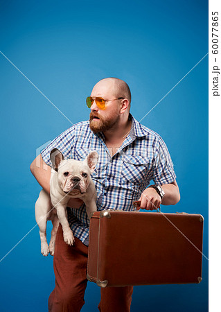 Bearded male in glasses with bulldog and luggage on empty blue background Bearded male in glasses with bulldog and luggage on empty blue background 60077865
