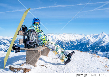 Photo from side of athlete man sitting on bench next to snowboard in ski resort. 60101630