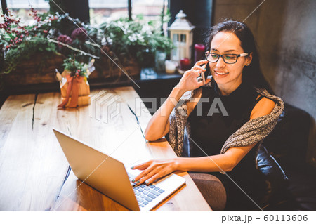 Beautiful young brunette woman inside cafe with christmas interior at wooden table by the window talking on phone and looking the laptop.In winter,she is dressed in a gray knitted sweater and glasses Beautiful young brunette woman inside cafe with christmas interior at wooden table by the window talking on phone and looking the laptop.In winter,she is dressed in a gray knitted sweater and glasses 60113606