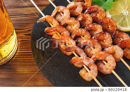 Shrimp fried on skewers with arugula, lemon on a black slate dish and beer in a glass on a wooden table 60120418