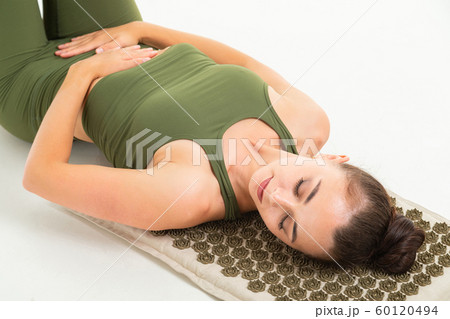 European brunette girl in a green sports suit meditates lying on a massage mat on a white background 60120494