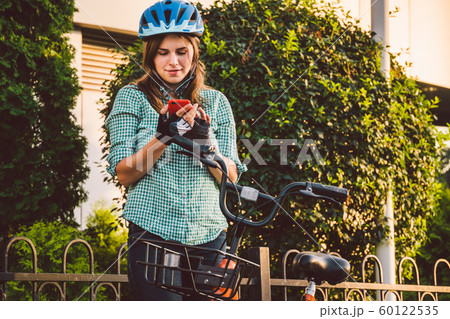 Portrait Of A Cheerful Girl Holding Mobile Phone. Happy smiling student using bike sharing app on smart phone outdoor. City lifestyle stylish hipster girl with bike using smartphone app in street Portrait Of A Cheerful Girl Holding Mobile Phone. Happy smiling student using bike sharing app on smart phone outdoor. City lifestyle stylish hipster girl with bike using smartphone app in street 60122535