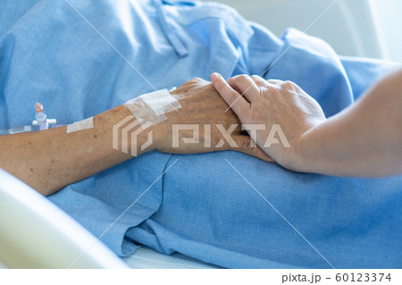 Caregiver holding elderly senior patient (ageing old adult person) hand in hospital bed or nursing hospice, geriatrician palliative home, while caretaker having  medical health care service 60123374