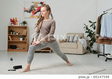 Young barefoot woman in activewear stretching legs while exercising on carpet 60147599