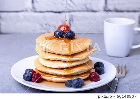 Pancakes with maple syrup, blueberries, cherries and a mini apple. Maple syrup pours on pancakes. A cup of coffee in the background. Traditional american pancakes. Close-up. 60148907