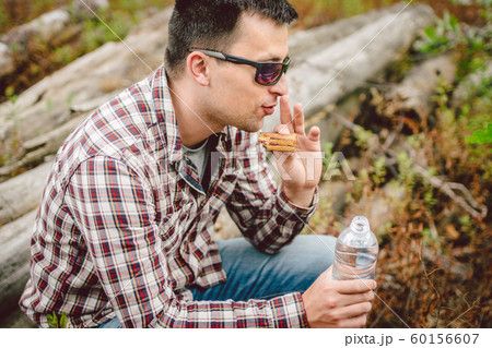 Fast food theme snack on nature. Caucasian man eating a sandwich and drinking water from a palette bottle in the forest. Tourist stopped for lunch in a wooded area 60156607