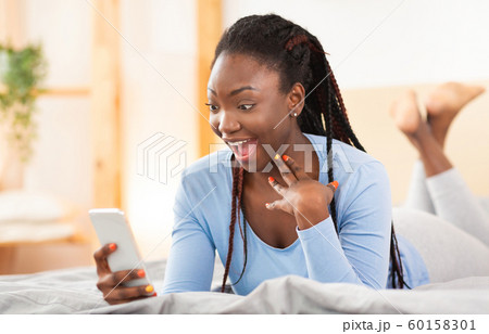 Excited Afro Woman Holding Smartphone Lying In Bed At Home Excited Afro Woman Holding Smartphone Lying In Bed At Home 60158301