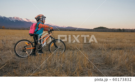 One caucasian children walk with bike in wheat field. Little girl walking black orange cycle on background of beautiful snowy mountains. Biker stand with backpack and helmet. 60173832