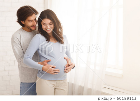 Loving man hugging his pregnant wife from behind standing near window Loving man hugging his pregnant wife from behind standing near window 60189113