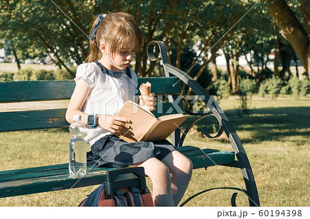 Portrait of schoolgirl 7 years old on a bench reading book, eating ice cream. Background city park 60194398