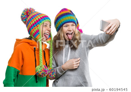 Winter portrait of two happy beautiful teenage girlfriends in knitted hats having fun with mobile phone, taking selfie, isolated on white background. People, youth and friendship concept Winter portrait of two happy beautiful teenage girlfriends in knitted hats having fun with mobile phone, taking selfie, isolated on white background. People, youth and friendship concept 60194545