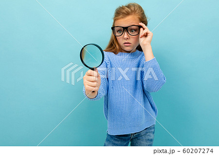 curious european girl in glasses looks through a magnifying glass on a light blue background curious european girl in glasses looks through a magnifying glass on a light blue background 60207274