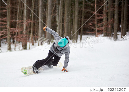 Male snowboarder in blue helmet, risky sliding on flat snow-covered road. Tree trunks and dark forest on background. 60241207
