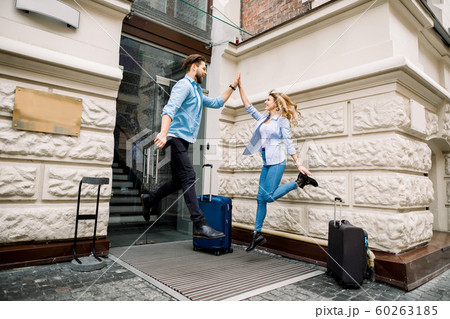 Young cheerful couple with suitcases jumping and giving five, having fun, before arriving to the hotel. Couple having fun outdoors, near the old city building Young cheerful couple with suitcases jumping and giving five, having fun, before arriving to the hotel. Couple having fun outdoors, near the old city building 60263185